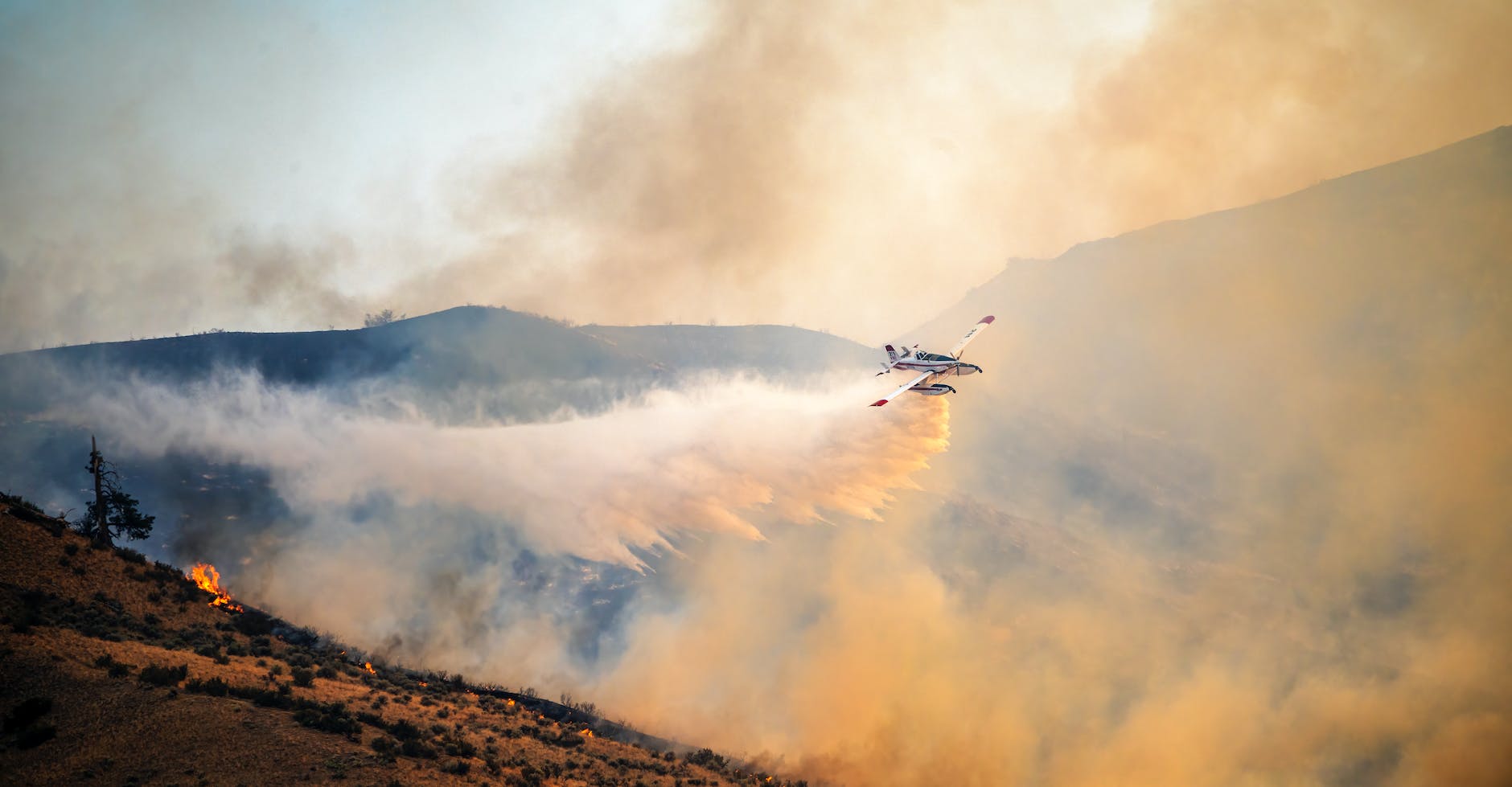 airplane flying over burning hilly terrain and spreading extinguishing powder
