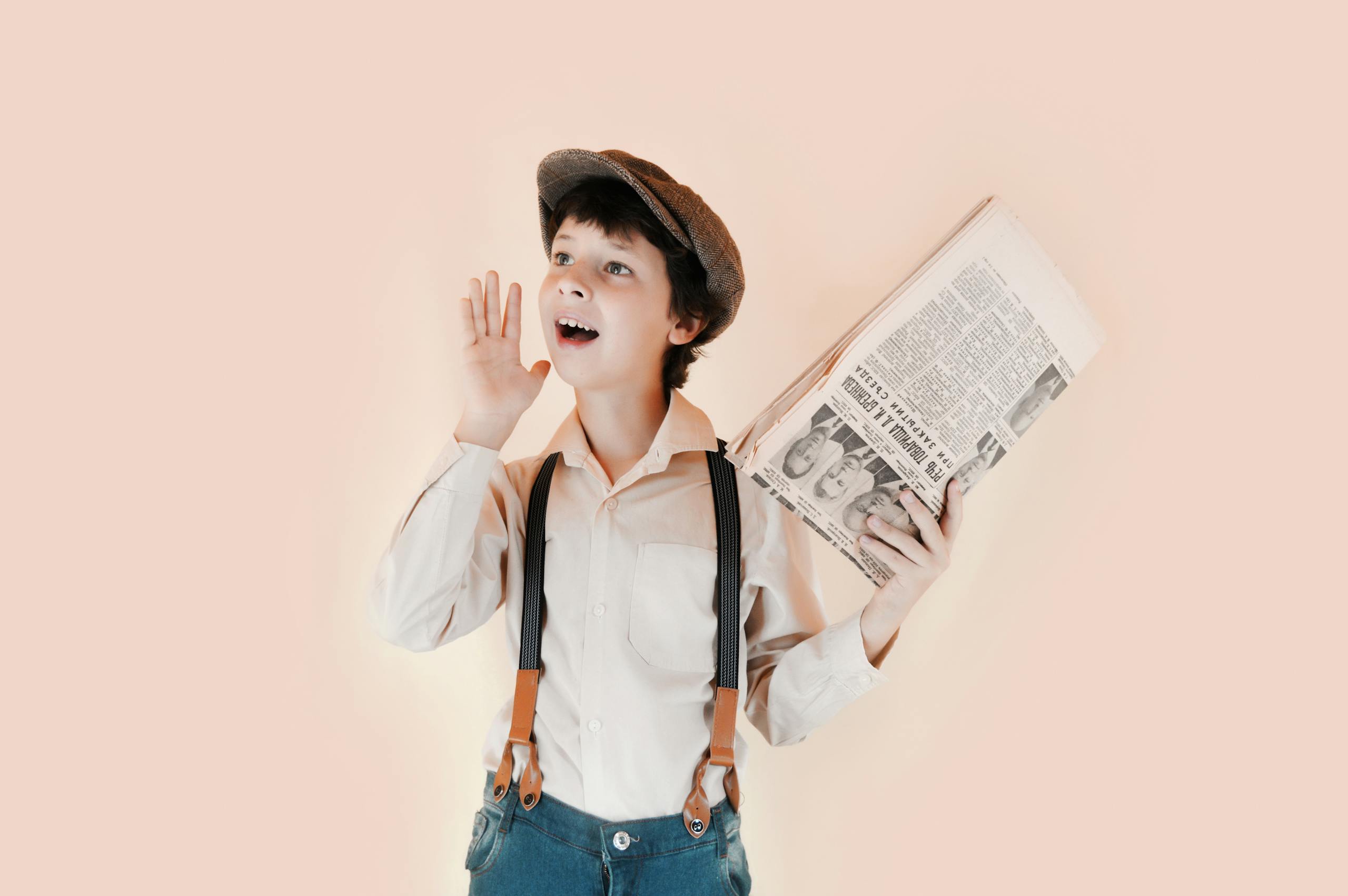 Positive boy in retro clothes holding old newspaper and standing with open mouth and raised arm against beige background in studio